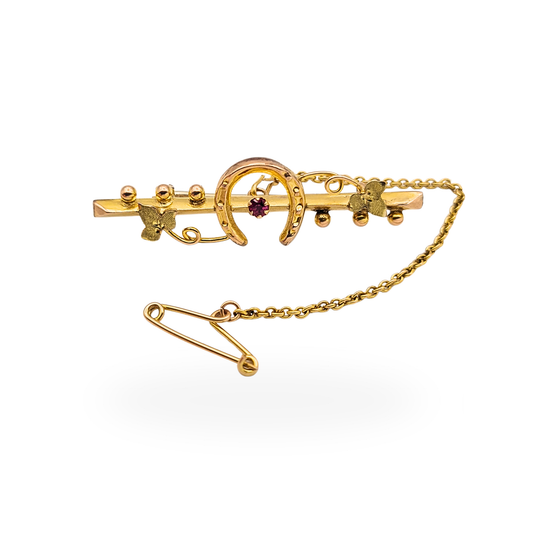 Edwardian 9ct gold horseshoe brooch measuring 37mm with central ruby detail, Birmingham 1906 hallmark, featuring bead and leaf decoration with safety chain and traditional stock pin fitting, lightweight hollow construction with antique finish